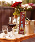 Rustic Cold Beer Served Here Metal Wall Sign Retro Vertical Tin Decor displayed on a wooden table with drinks and flowers.