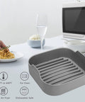 Grey air fryer silicone pot 8.1 inch basket pan with dual handles for 6.5 Qt square fryers next to a pasta plate on a table.