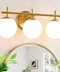 Next Level Three Light Bathroom Vanity Lights In Brushed Gold Finish With Milky White Glass Globe Shades Modern Wall Sconce Over Mirror viewed above a mirror with greenery.