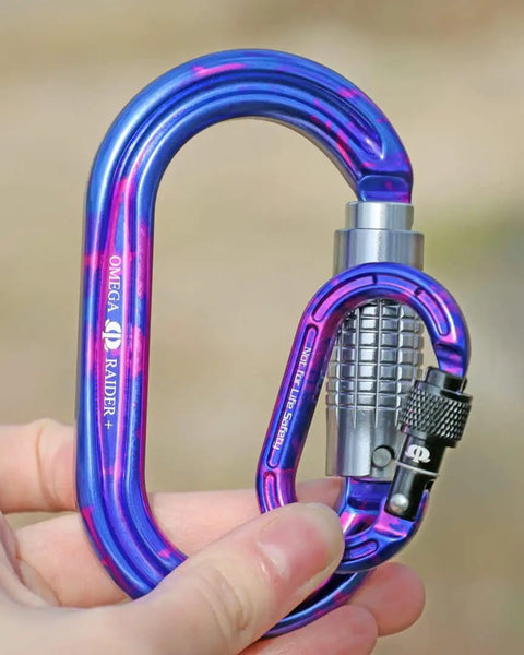 Person holding blue Omega Raider screw-lock carabiner with black locking mechanism, outdoor setting.