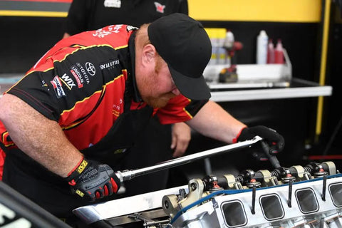 Mechanic wearing heavy duty shop apron working on an engine, showcasing professional use of the Heavy Duty Shop Apron Built Tough With Liquid Resistant Fabric And A Boatload Of Tool Pockets Center Chest Pocket Machine Washable Adjustable Straps One Size Fits All Black