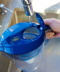Filling a blue water pitcher with Pure Replacement Filter For Water Pitcher That Removes Fluoride Lead Forever Chemicals Heavy Metals And Industrial Pollutants Delivers Clean Great Tasting Water 100 Gallon Capacity Easy Swap attached.