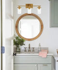 Brushed Brass 3 Light Vanity Light With Clear Glass Shades in bathroom setting, above round mirror, sleek urban vintage vibe
