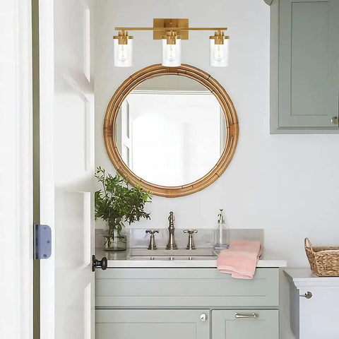 Brushed Brass 3 Light Vanity Light With Clear Glass Shades in bathroom setting, above round mirror, sleek urban vintage vibe