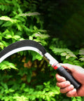 Hand holding Two Pack Manganese Steel Weeding Sickle Set For Easy Yard Work, Weed Cutting And Harvesting in front of green foliage.