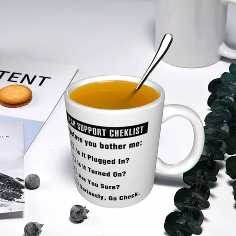 11oz Funny Tech Support Checklist Coffee Mug for Help Desk Heroes filled with tea and spoon, surrounded by books and snacks.