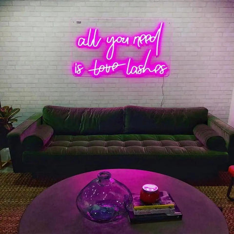 Pink 'All You Need Is Love Lashes' neon sign above a green couch in a living room setting