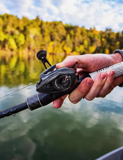 Person holding Torrent Lite Ultra Light Baitcasting Reel With Carbon Body, 5.5 Oz Lightweight Build, Black, by a lake.