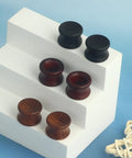 Three Pairs Organic Wood Ear Plugs Set With Double Flared Ear Tunnels Saddle Fit Wooden Gauges Five Eighths Inch 16mm Natural Black Brown Tones Perfect For Punk Piercing And Everyday Wear displayed on a white stand