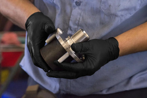 Black nitrile gloves on hands, holding a metal mechanical component with bolts