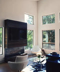 Modern living room with large windows, featuring a wood accent wall, TV, white chairs, and glass coffee table.