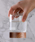 Hand holding a Slick High Borosilicate Glass Storage Jar With Acacia Wood Lids filled with water on a marble surface.