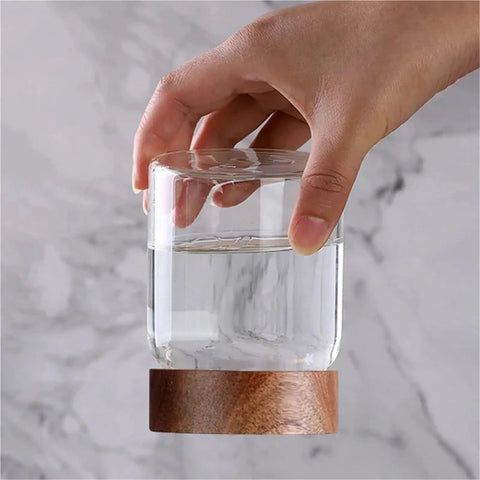 Hand holding a Slick High Borosilicate Glass Storage Jar With Acacia Wood Lids filled with water on a marble surface.