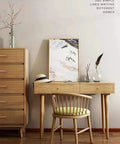Minimalist wooden desk setup with chair in a cozy room, next to a tall chest, featuring natural decor accents and a framed artwork.