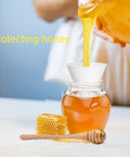 Glass jar with honey poured through a paper funnel into the jar, hexagonal honeycomb and dipper nearby.