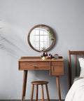 Modern wooden vanity with round mirror and stool, next to a bed in a minimalist bedroom, against grey wall.
