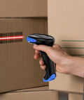 Hand holding a 2D Bluetooth Barcode Scanner with 3 in 1 Connectivity scanning a barcode on a cardboard box in a warehouse setting.