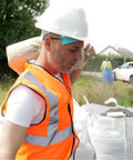 Construction worker in hard hat and orange vest carrying bag on shoulder at a job site with containers in background.