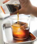 A hand pouring cold brew coffee over ice in a glass, showcasing the rich color of the brew made using Cold Brew Filter Bags 100 Pack For Home Use Disposable Coffee Brewing Bags Measuring 11.25 x 13 Inches Strong Enough To Hold Up To 10 Lbs Ground Coffee For Bold, Smooth Cold Brew At Home.