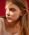 Close-up of woman wearing a small silver nose stud and long dangling earrings with red accents, against a red background.