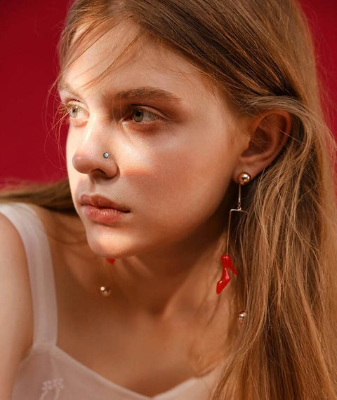 Close-up of woman wearing a small silver nose stud and long dangling earrings with red accents, against a red background.