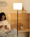 Floor lamp with black metal pole and white fabric shade, 60-inch tall, shown beside a person sitting on a bed.