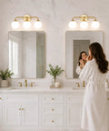 Dope Mid Century 3 Light Vanity Fixture With Smooth Brushed Gold Finish And Soft Milky White Glass Ball Shades For Sick Bedroom Bathroom Mirror Vibe, mounted over double mirrors in a modern bathroom.