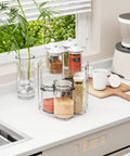 Totally Slick No Hassle 360 Spin Vanity Organizer Two Tier Clear Acrylic Rotating Storage For Perfume And Lotion Lazy Susan Beauty Stand Keeps Skincare Fully Within Reach On Any Desk displayed on a kitchen countertop with jars and a plant.
