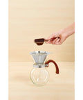 Next Level Pour Over Coffee Maker With Heat Resistant Borosilicate Glass Carafe And Comfortable Bamboo Handle Plus Durable Stainless Steel Filter For Killer Black Coffee And Straight Up Smooth Sips Daily with coffee grounds being added.
