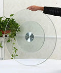 Hand holding a Heavy Duty Tempered Glass Lazy Susan Serving Platter with a sleek design near a plant.