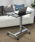 Smooth Roll Mobile Standing Desk With Height Adjustable Pneumatic Lift Sleek Black Aluminum Frame Ergonomic Rolling Workstation beside a couch, laptop and mouse on black surface.