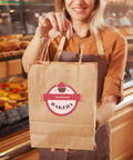 Person holding Kraft Paper Gift Bags With Handles 8x4.25x10.5 Inches Bulk Pack Of 100 Brown Recycled Bags in bakery setting