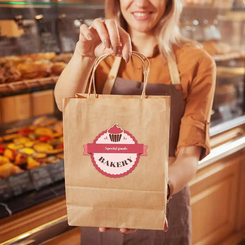 Person holding Kraft Paper Gift Bags With Handles 8x4.25x10.5 Inches Bulk Pack Of 100 Brown Recycled Bags in bakery setting