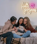 Hello Gorgeous Neon Sign For Girls Bedroom Wall Decor LED Light With Warm White Glow Perfect For Birthday Parties Weddings And Gifts 16.7x12.3 Inches Great Gift Idea illuminated above three people sitting on a couch with a dog.