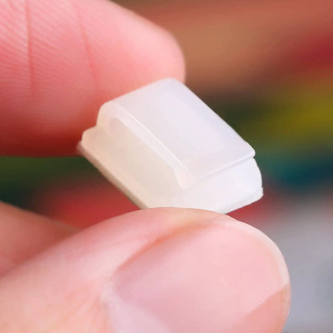 Close-up of a white self-adhesive cable clip held between fingers, showing detail for Adhesive Cable Clips 100 Pack Go To Cable Management Self Adhesive Wire Clips Cord Holders Small White For Car Outdoor Wall String Lights Desk Home Office Organization.