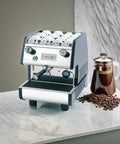 Hardcore Pro Programmable Espresso Maker Featuring Digital Touch Controls And Precision Volumetric Dosing For Dead Smooth Shots Heavy Duty Brass Group Head Matte Black Finish on marble countertop with coffee beans and a French press.