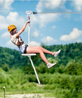 Person riding zipline with Extra Long Zipline Spring Brake For Backyard Kits, over green landscape.