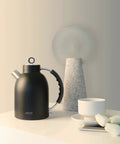Retro Glass Electric Kettle 1.5 Liters Tea Boiler With Bpa Safe Cordless Setup Straight Up Plastic Free Build And Auto Shut Off Protection In Sleek Black next to a white teacup and flowers.