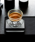 Glass of espresso on a black table next to Espresso Tamping Mat With Portafilter Dock, Food Grade Silicone Coffee Tamper Mat For Baristas.