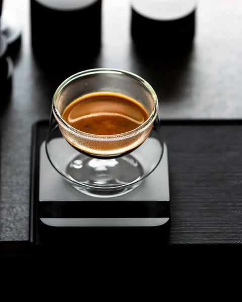 Glass of espresso on a black table next to Espresso Tamping Mat With Portafilter Dock, Food Grade Silicone Coffee Tamper Mat For Baristas.