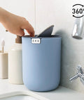 Person placing item in Ultra Compact 1.5L Mini Tabletop Trash Can With Swing Lid For Bathroom Vanity Or Desk Space Saving 4.7 Inch Diameter 6.5 Inch Tall Small Garbage Bin For Office Countertop on sink