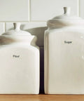 Two white ceramic storage canisters labeled 'Flour' and 'Sugar' on a wooden surface, suitable for kitchen organization.