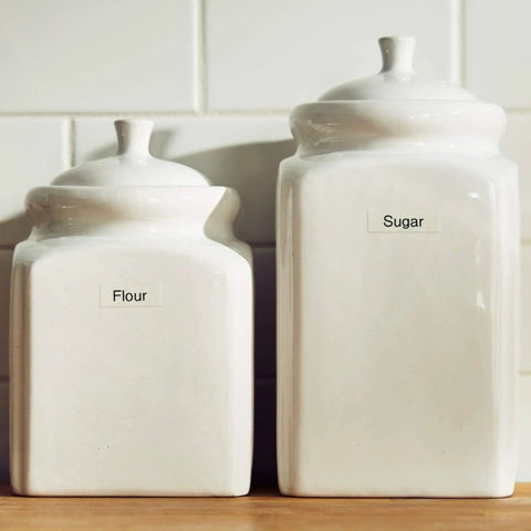Two white ceramic storage canisters labeled 'Flour' and 'Sugar' on a wooden surface, suitable for kitchen organization.