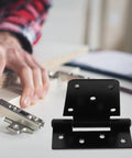 Heavy Duty Furniture Hinges Set In Black Carbon Steel With Screws, shown on a table with a person assembling furniture nearby.