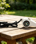 3-In-1 Survival Whistle With Built-In Compass And Thermometer on wooden table, white color, with attached black lanyard.