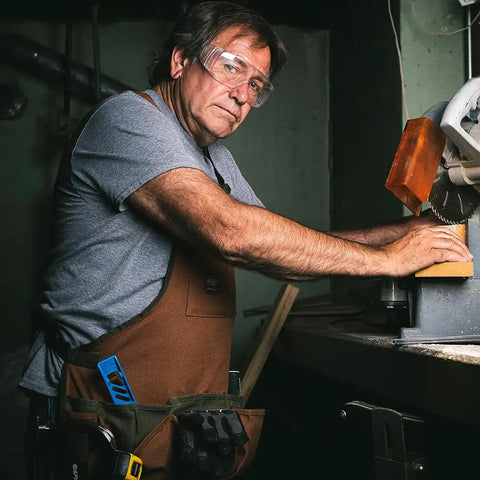 Man using a saw wearing Heavy Duty Canvas Tool Bag For Men With 16 Pockets, Padded Adjustable Shoulder Straps, Brown.