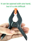 Front view of a 4.5-inch black plastic spring clamp with orange jaw, held by hand.