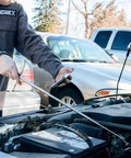 Mechanic using a telescoping antenna on a car battery outside in a parking lot with other vehicles in the background.