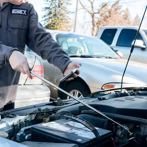 Mechanic using a telescoping antenna on a car battery outside in a parking lot with other vehicles in the background.
