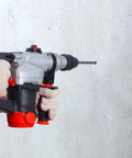 Side view of an SDS Plus hammer drill with a masonry bit held by gloved hands against a concrete wall.